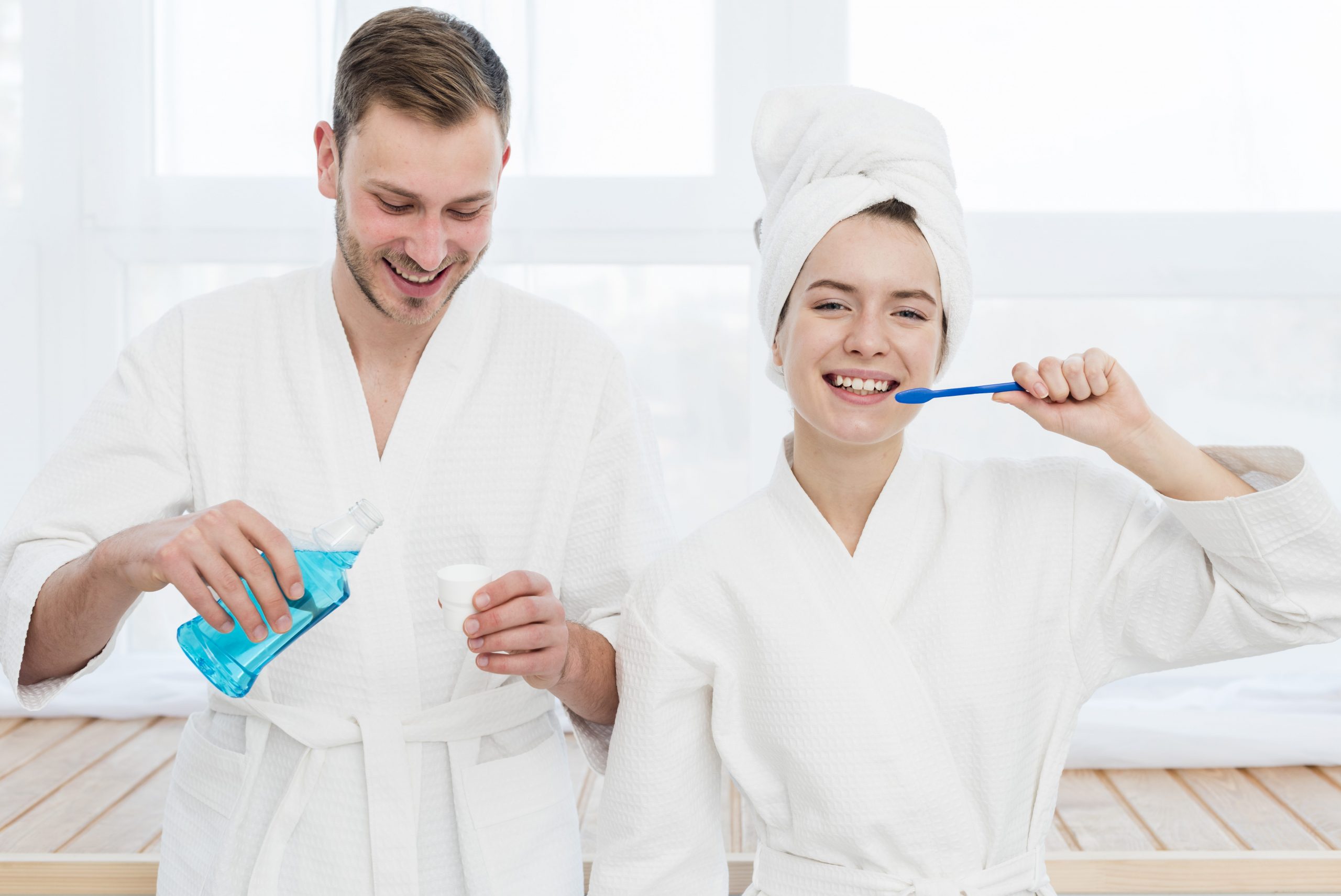 A couple in the bathroom, the man is pouring mouthwash on the lid and the woman is brushing her teeth.