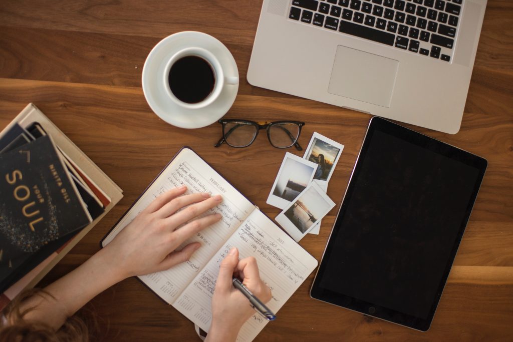 A woman's hand writing in a journal over a desk with a notebook, glasses, a cup of coffee, some books and pictures.